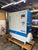 A large industrial air compressor unit with a white and blue cabinet and a white cylindrical tank, sitting on a wooden pallet in a workshop or garage setting. Visible branding includes COMBI on the side and ALMIG on the front panel.