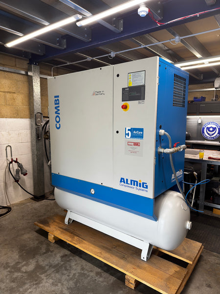 A large industrial air compressor unit with a white and blue cabinet and a white cylindrical tank, sitting on a wooden pallet in a workshop or garage setting. Visible branding includes COMBI on the side and ALMIG on the front panel.