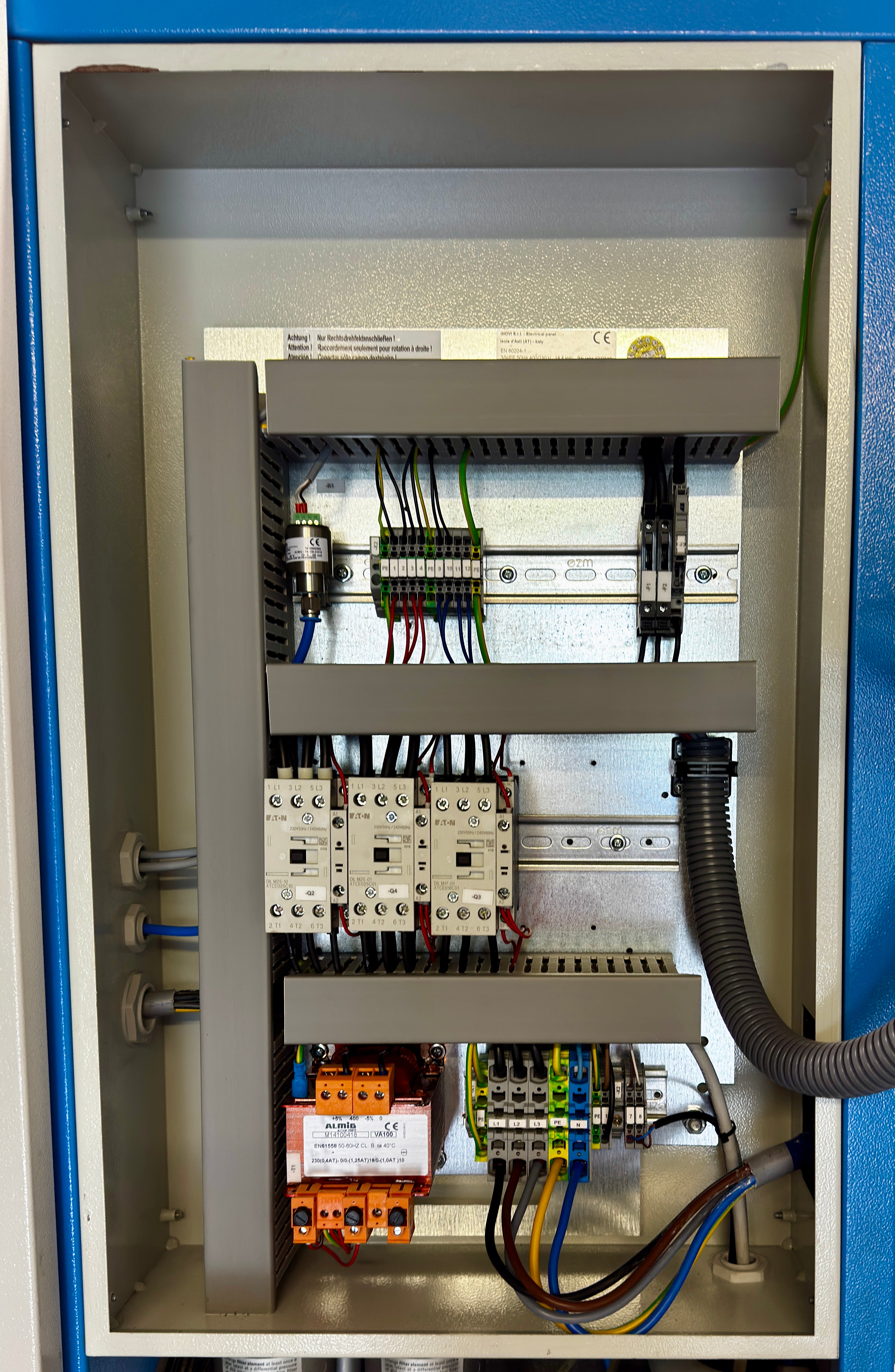 Open industrial electrical control panel showing internal components including contactors, terminal blocks, wiring, and a black flexible conduit inside a metal cabinet.