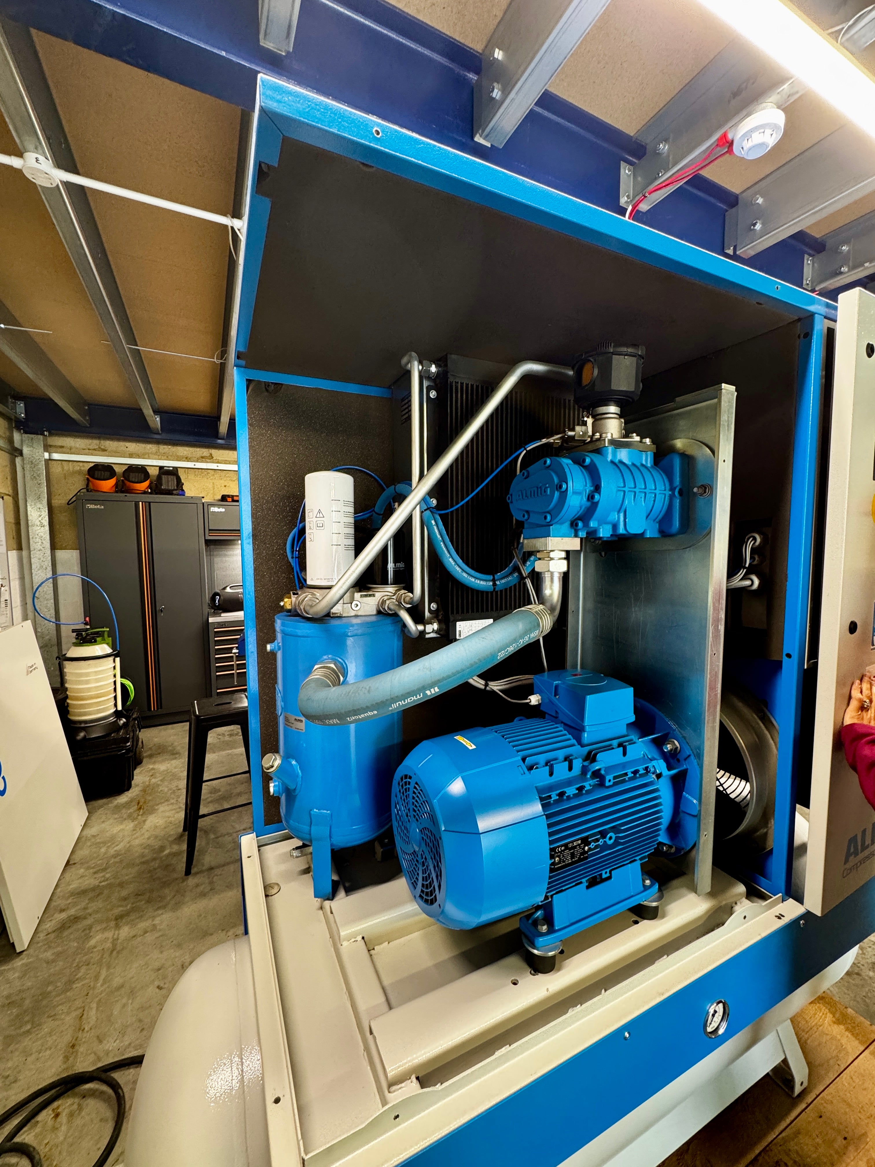 A large blue and beige industrial machine cabinet with an open front panel, showing a blue electric motor, pumps, and piping inside a workshop or factory setting.