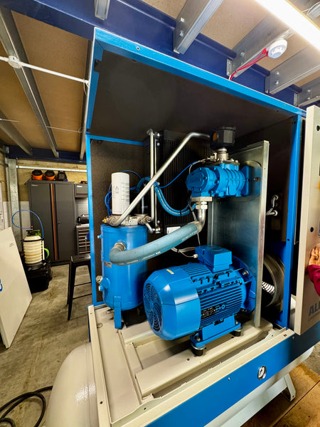 A large blue and beige industrial machine cabinet with an open front panel, showing a blue electric motor, pumps, and piping inside a workshop or factory setting.