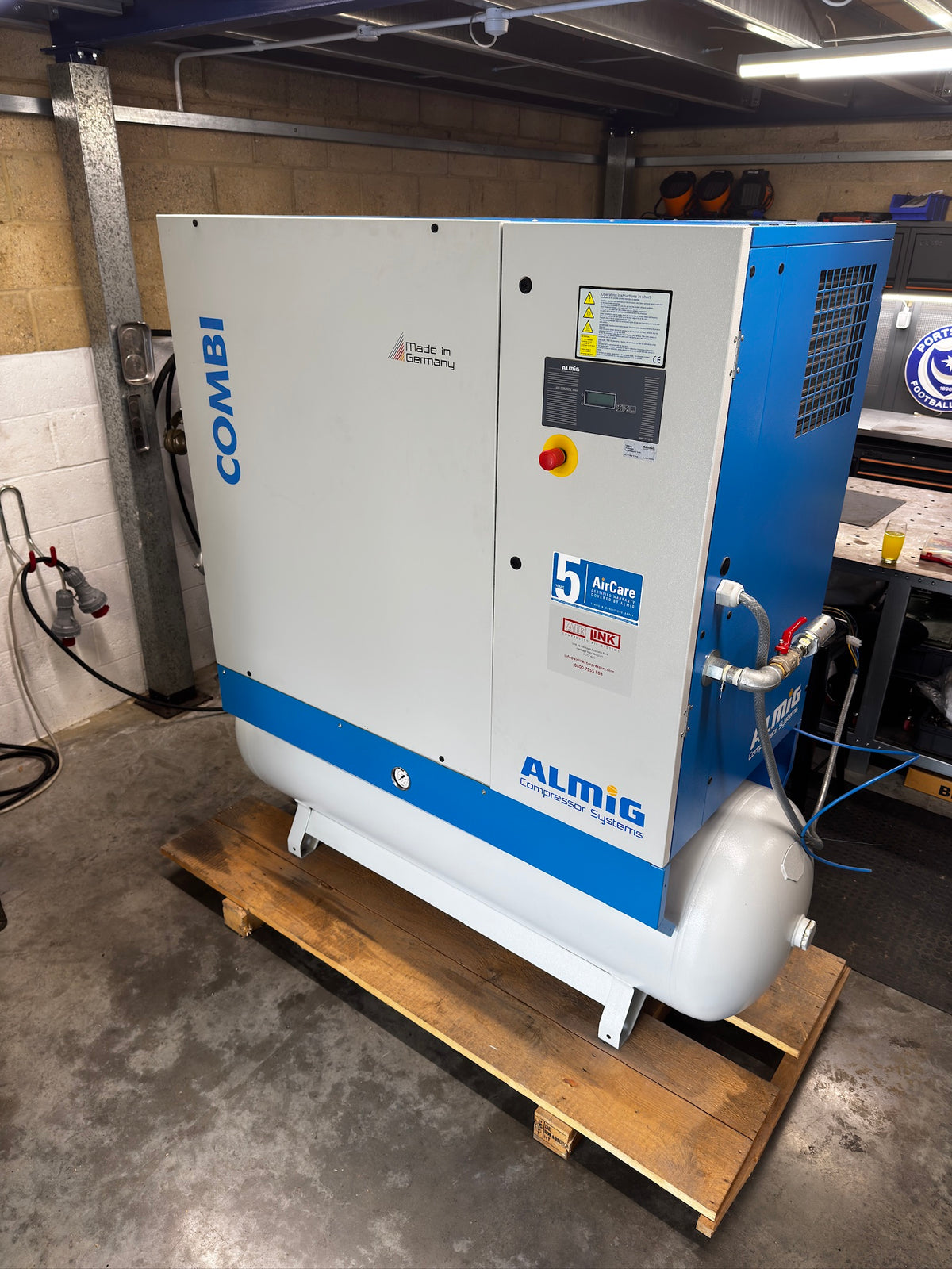 A large Combi ALMIG industrial air compressor with a white and blue cabinet, mounted on a wooden pallet in a workshop environment.