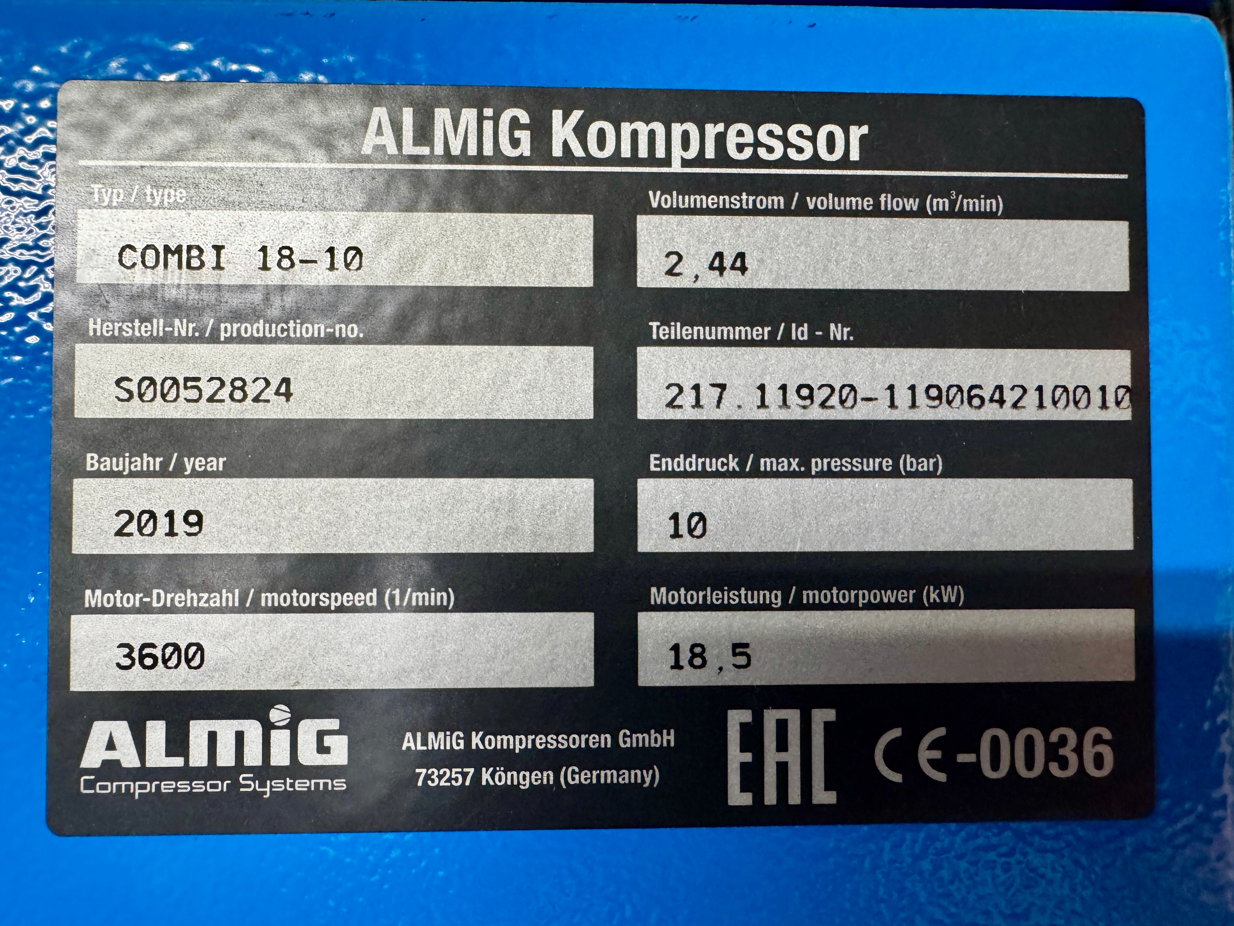A close-up of a black ALMiG compressor label with white text and fillable fields, photographed against a bright blue surface. Visible text includes 'ALMiG Kompressor', model information, serial number, year, motor speed, pressure, and EAC certification marks.
