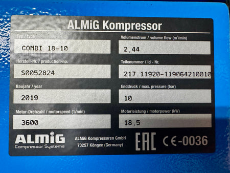 A close-up of a black ALMiG compressor label with white text and fillable fields, photographed against a bright blue surface. Visible text includes 'ALMiG Kompressor', model information, serial number, year, motor speed, pressure, and EAC certification marks.