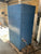 A large blue industrial machine or cabinet photographed in a workshop or utility space, with pipes, cords, and concrete floor visible around it.