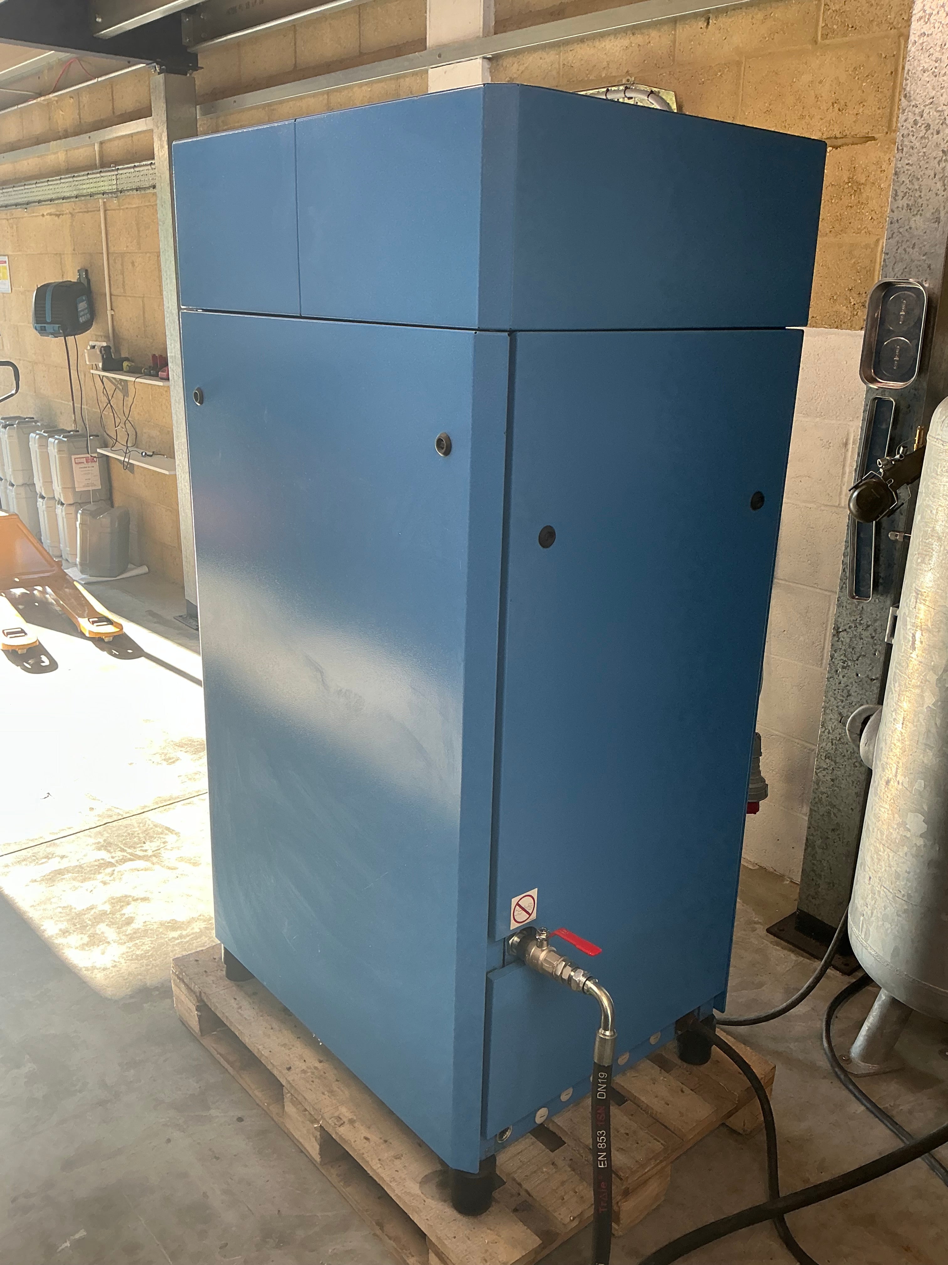 A large blue industrial machine or cabinet photographed in a workshop or utility space, with pipes, cords, and concrete floor visible around it.