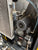 Close-up of a mechanical industrial compressor or air pump unit with a large metal radiator, cylindrical motor housing, belts, hoses, and piping.