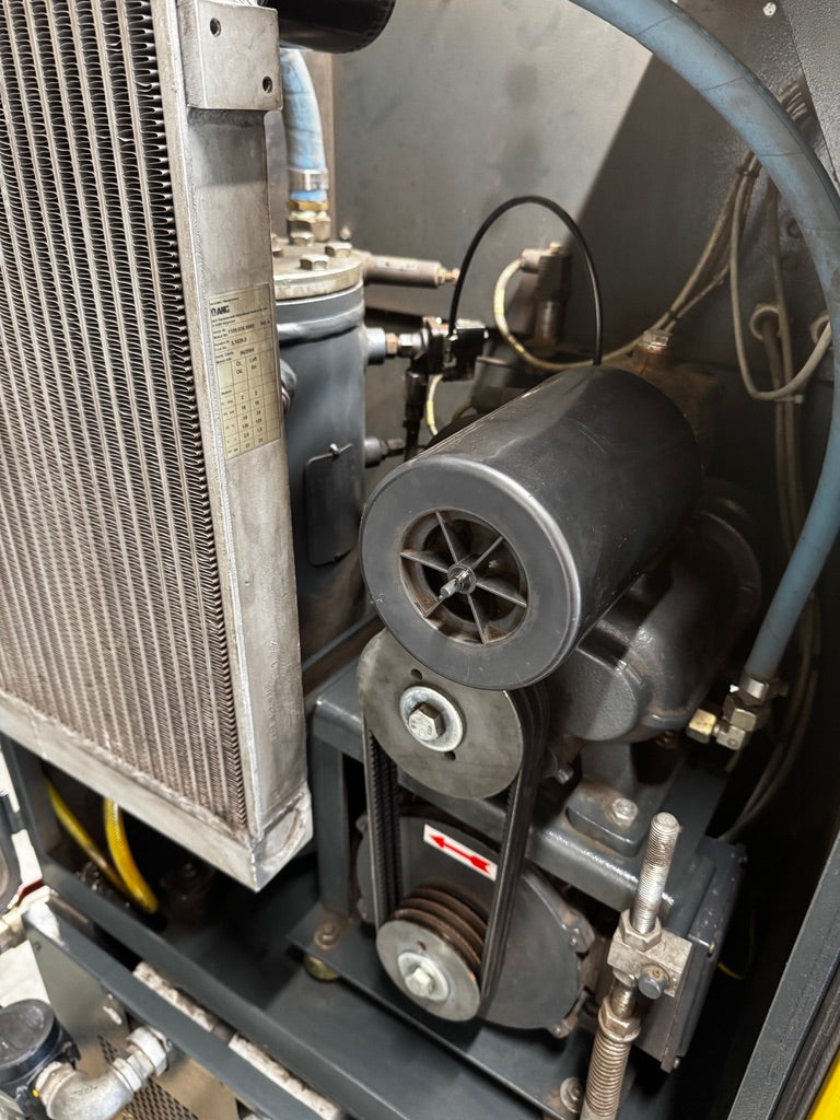 Close-up of a mechanical industrial compressor or air pump unit with a large metal radiator, cylindrical motor housing, belts, hoses, and piping.