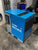 A blue and gray industrial air compressor by MSA/IMARK, photographed in a workshop or garage setting with concrete floors and storage containers in the background.