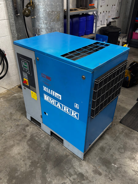 A blue and gray industrial air compressor by MSA/IMARK, photographed in a workshop or garage setting with concrete floors and storage containers in the background.