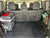 A black diamond-plate cargo liner mat installed in the trunk of a vehicle, shown with the rear seats folded down. A gray cooler bag with a red lid and a green rope are placed on the left, and a green storage bin with a red bottle is on the right.