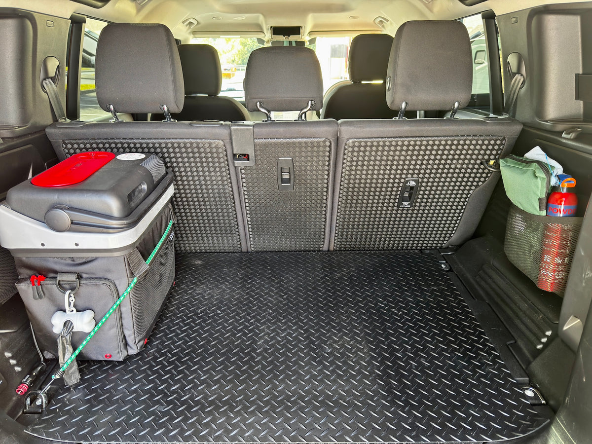 A black diamond-plate cargo liner mat installed in the trunk of a vehicle, shown with the rear seats folded down. A gray cooler bag with a red lid and a green rope are placed on the left, and a green storage bin with a red bottle is on the right.
