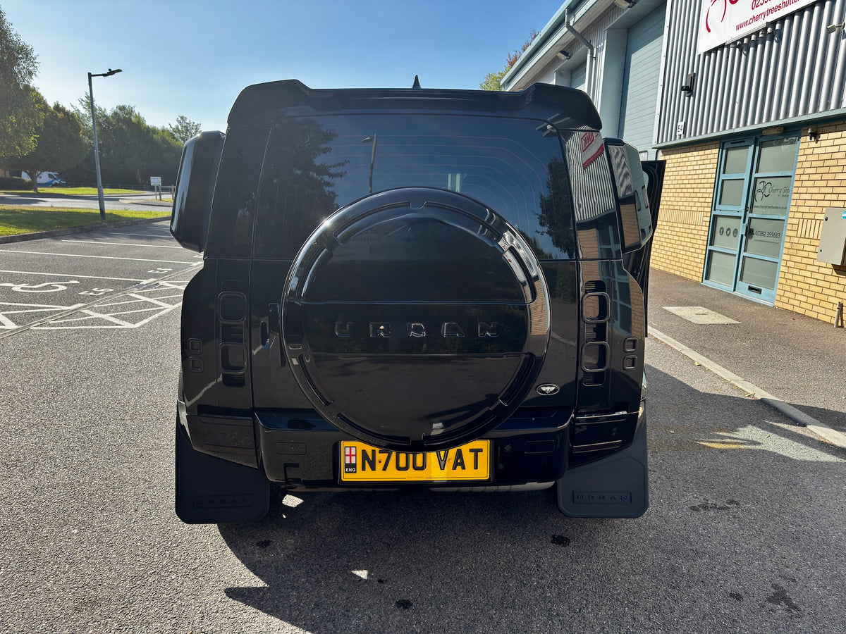 A black SUV shown from the rear, parked on an asphalt lot next to a building. The vehicle features a circular spare tire cover with the word 'URBAN' embossed in the center and a yellow license plate reading 'N700 VAT'.