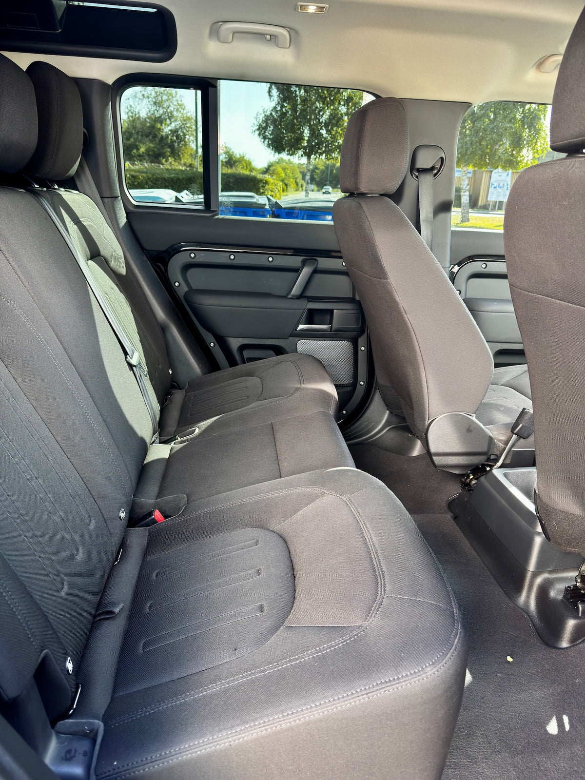 Rear passenger compartment of a vehicle showing the back seat, front seats, door panel, and window view with trees outside.