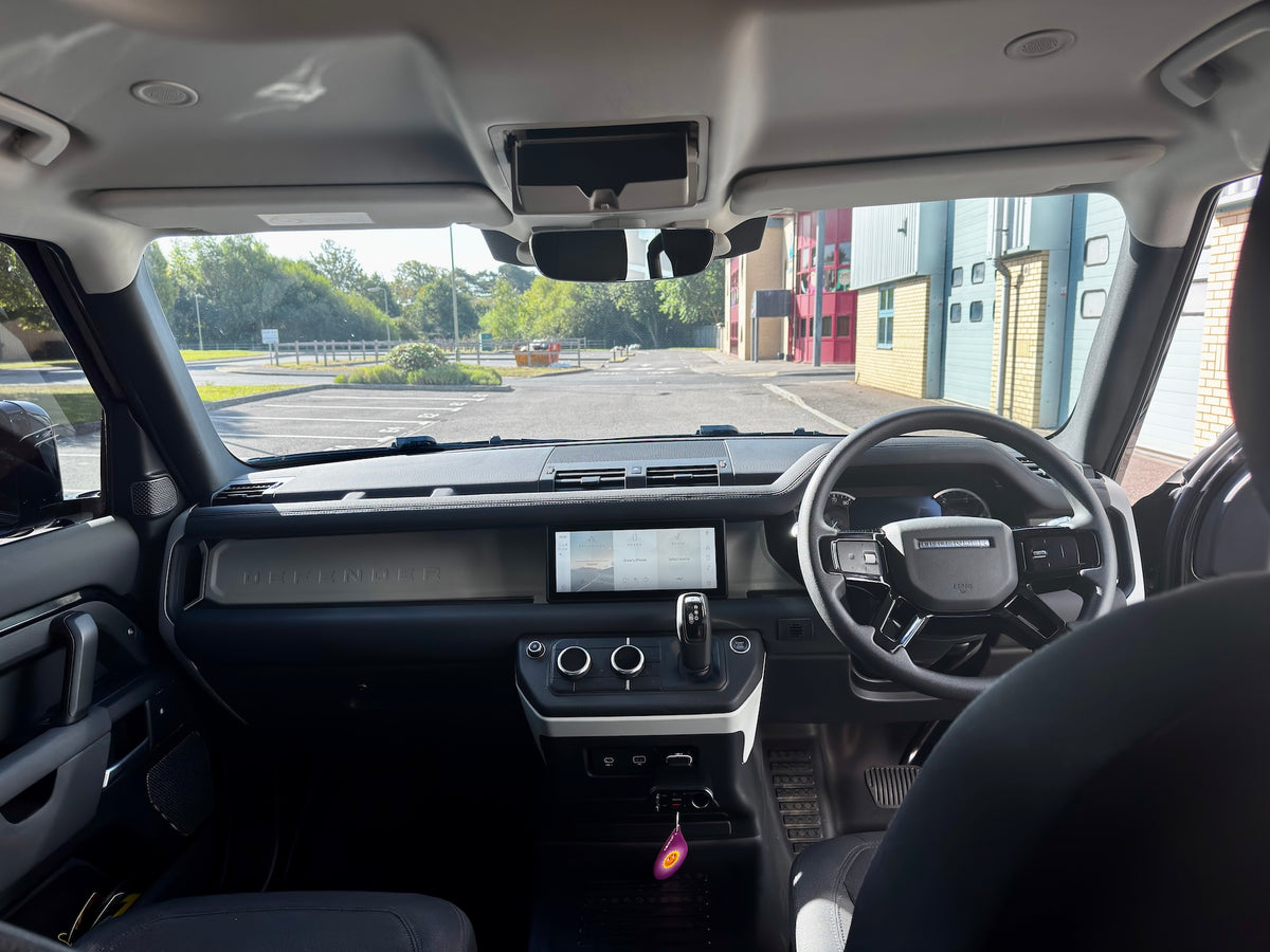 The interior of a modern SUV or Land Rover vehicle, showing the dashboard, steering wheel, center console, and headliner, with a view of an outdoor parking lot through the windshield.