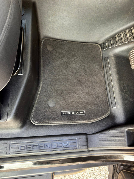 A black car floor mat with a vertical 'URANI' logo, installed in the footwell of a vehicle with a Defender door sill visible.