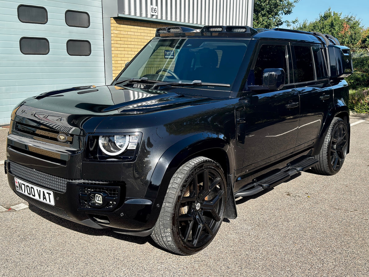 A black custom Land Rover Defender SUV parked on an asphalt driveway in front of a garage and brick building.