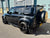 A black Land Rover Defender SUV parked outdoors in front of a light blue corrugated metal building. The vehicle is shown from a rear three-quarter angle, featuring black wheels, a spare tire mounted on the rear door, roof rails, and a yellow license plate.