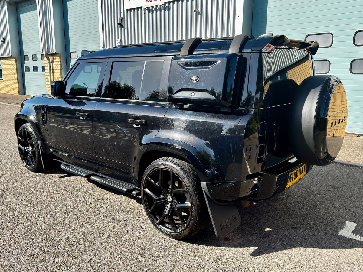 A black Land Rover Defender SUV parked outdoors in front of a light blue corrugated metal building. The vehicle is shown from a rear three-quarter angle, featuring black wheels, a spare tire mounted on the rear door, roof rails, and a yellow license plate.