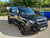 A black Land Rover Defender SUV parked outdoors on a paved surface, with trees and bins in the background. The front license plate reads N700 V17.
