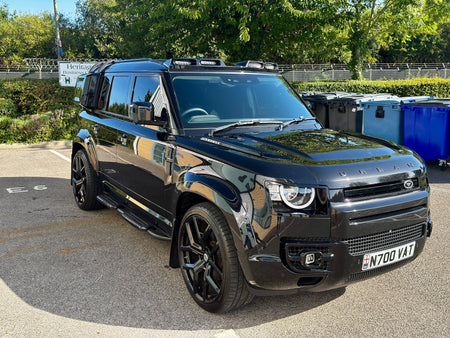 A black Land Rover Defender SUV parked outdoors on a paved surface, with trees and bins in the background. The front license plate reads N700 V17.