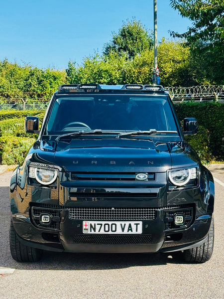 A black Land Rover Defender Urban SUV parked outdoors on a paved surface, facing forward. The vehicle features a chrome front grille, a license plate reading N700 VAT, and a roof rack with lights.
