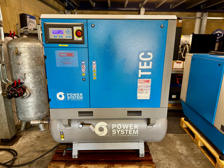 A blue and gray industrial air compressor by Power System TEC, photographed in a workshop or warehouse setting with other machinery visible in the background.