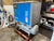 A large industrial air compressor from Power Systems TEC, featuring a blue and gray cabinet and a gray air tank, sitting on a wooden pallet in a workshop environment.